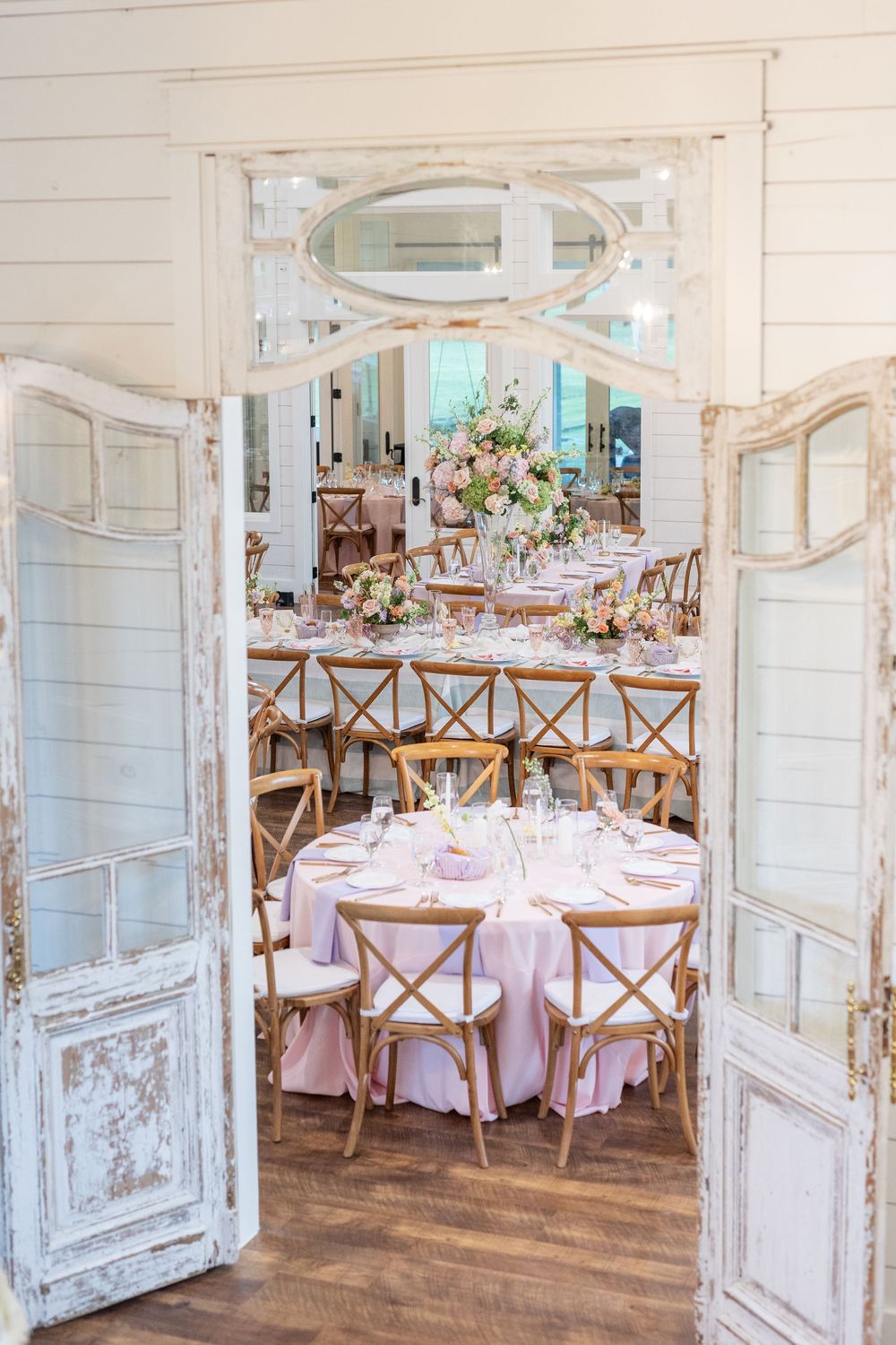 Wedding reception hall viewed through vintage distressed doors with pink linen tables and wooden chairs.