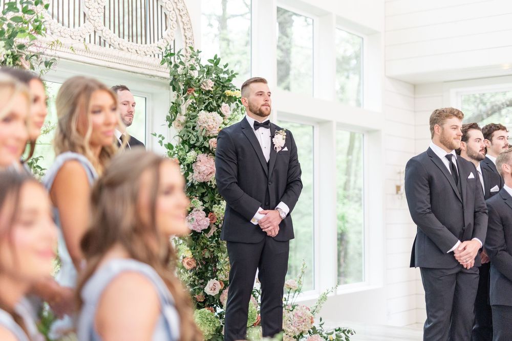 Groom in black tuxedo stands at altar awaiting bride during elegant indoor wedding ceremony.