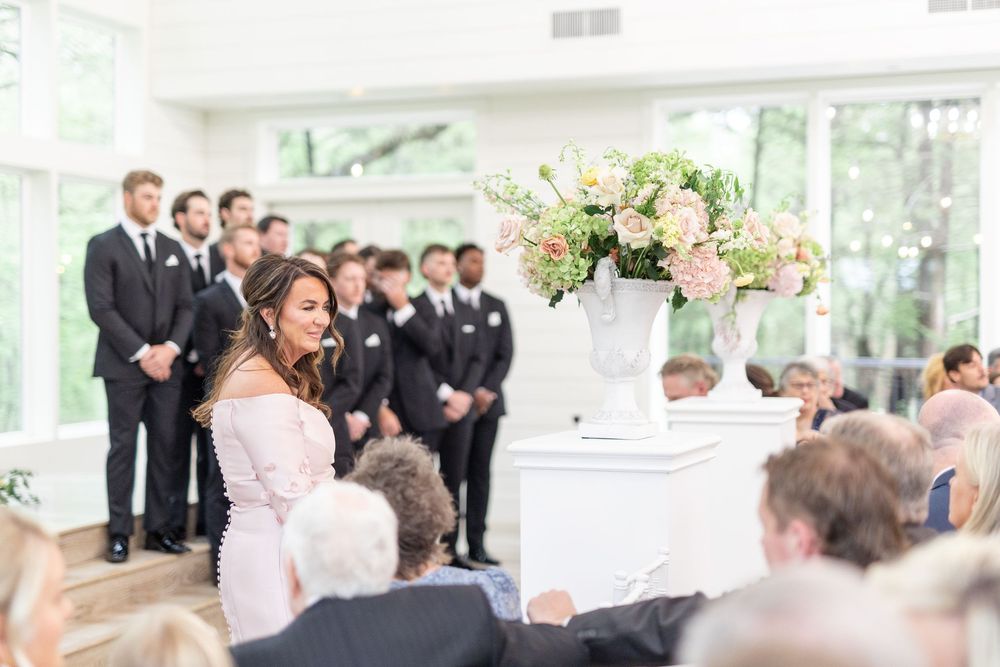 Woman in pink off-shoulder dress walks down aisle past groomsmen during bright indoor wedding ceremony.
