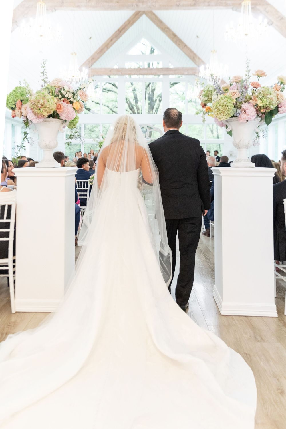 Bride in white gown with long veil walks down aisle escorted by father in black suit.