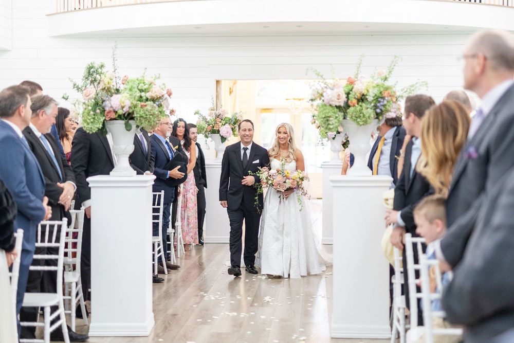 Bride carrying pink bouquet walks down floral-decorated aisle with escort during indoor wedding ceremony.