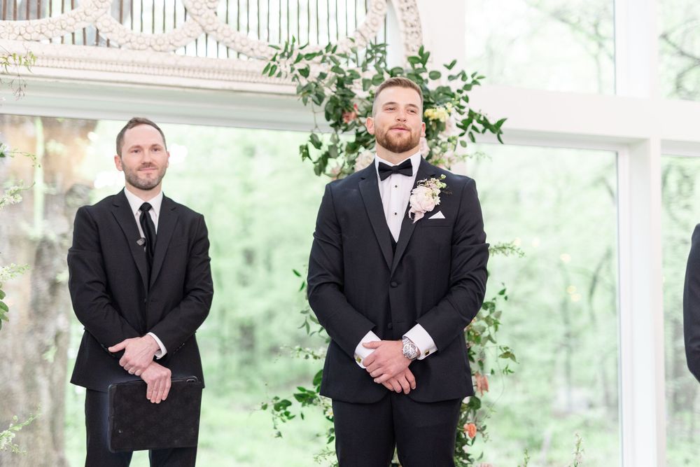 Groom in black tuxedo with floral boutonniere stands at altar with best man during wedding ceremony.