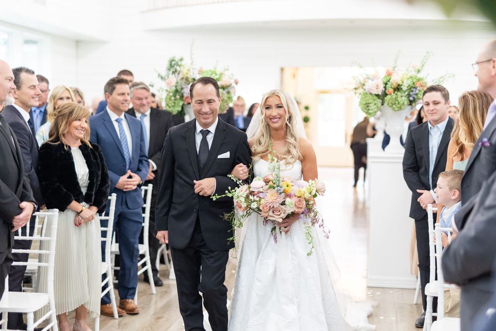 Smiling blonde bride in white gown carries colorful bouquet while walking down aisle with escort.