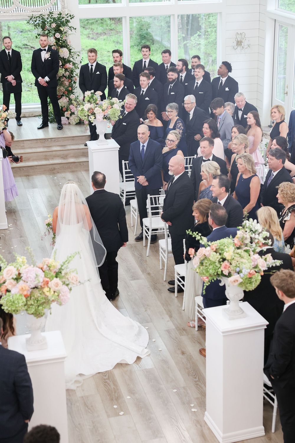 Aerial view of bride with long veil walking down aisle with guests standing in bright chapel.