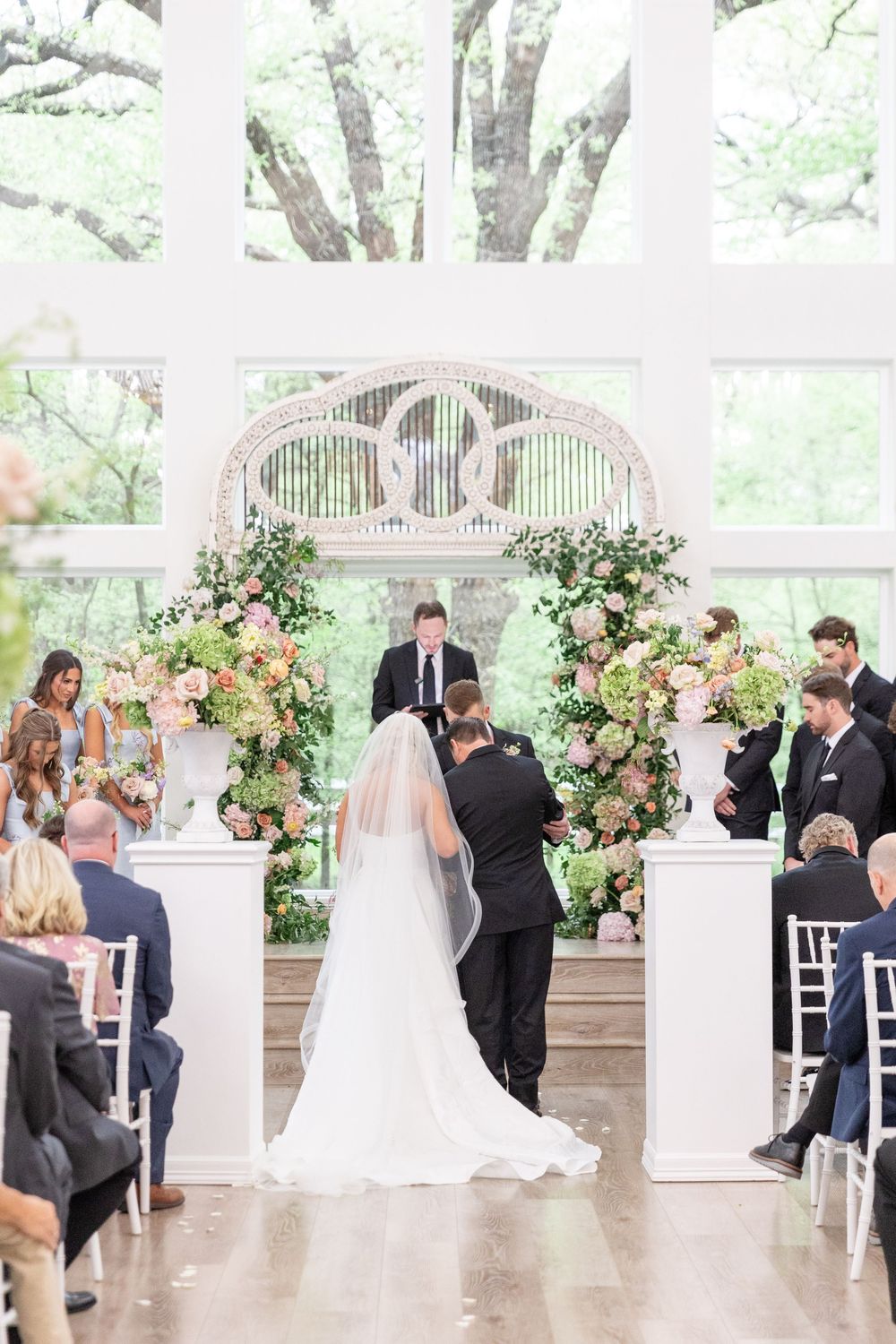 Bride and groom stand at floral arch altar during wedding ceremony in sunlit chapel.