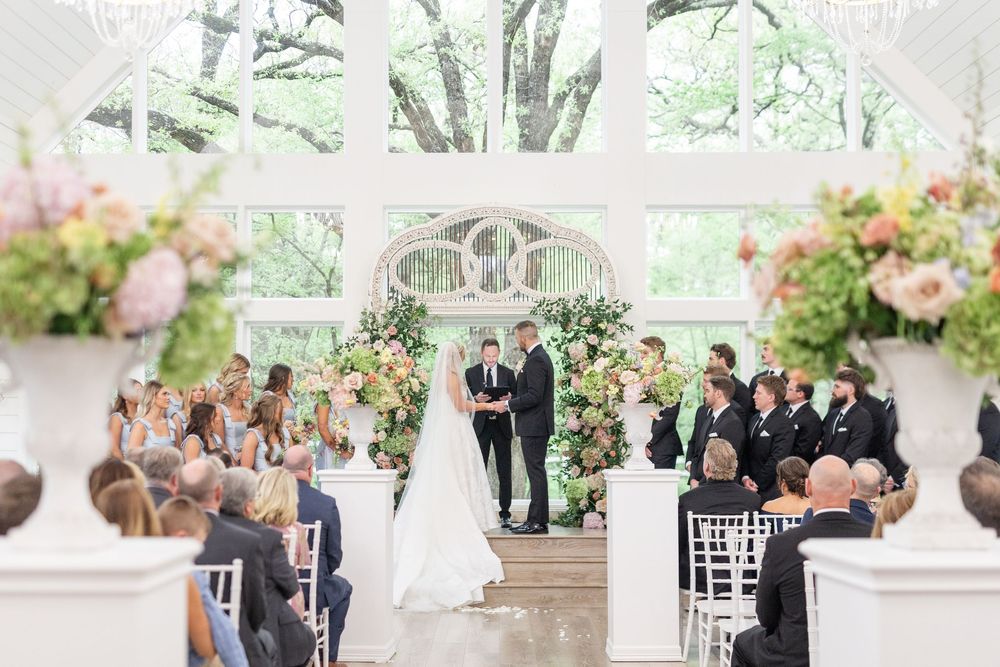 Wide shot of elegant wedding ceremony with couple at floral altar in bright windowed chapel.