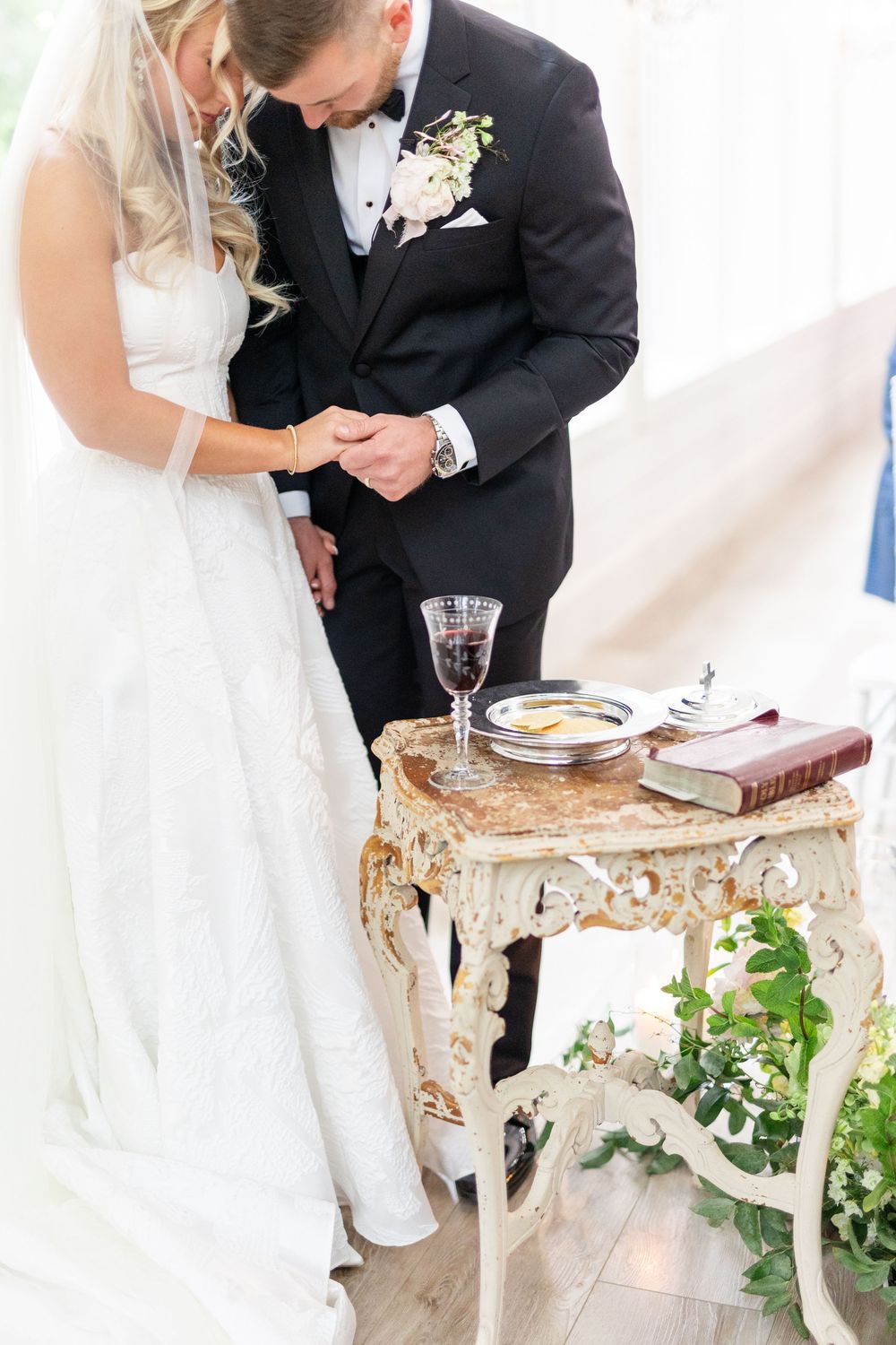 Bride and groom bow heads in prayer at ornate table with communion elements during ceremony.