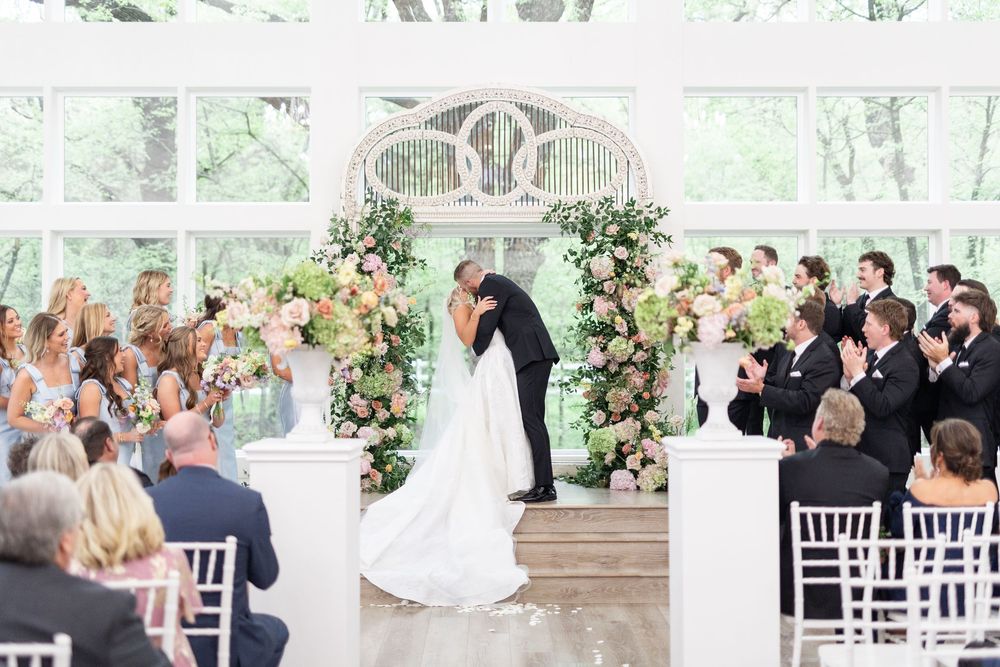 Bride and groom share first kiss at elegant indoor wedding ceremony with floral arch and white chairs.
