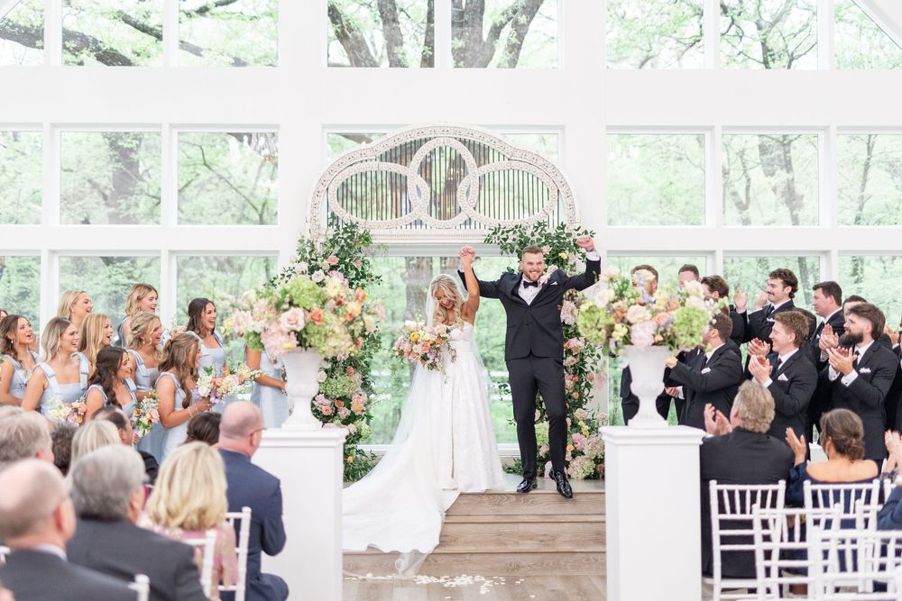 Bride and groom celebrate with raised arms at indoor wedding ceremony surrounded by floral arrangements.
