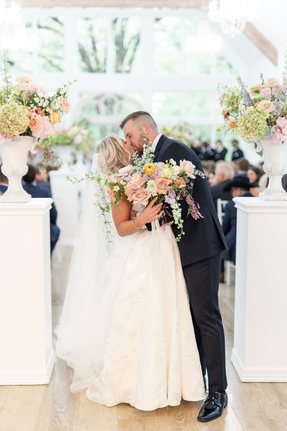 Bride and groom share a kiss at altar with colorful pastel floral arrangements in white urns.