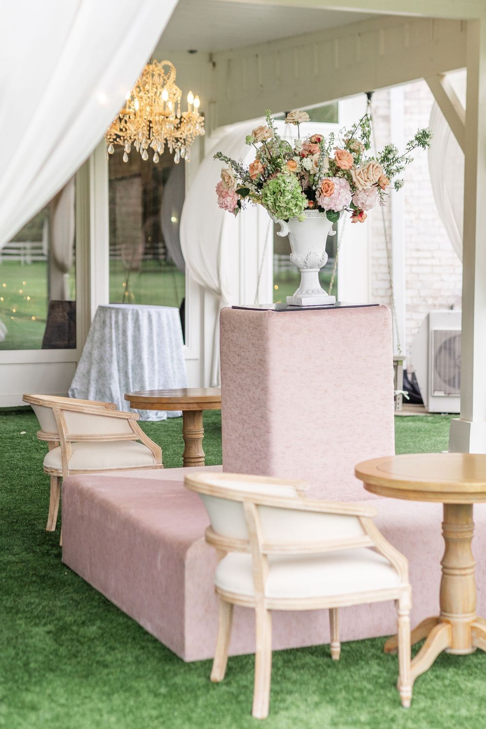 Elegant outdoor wedding lounge with pink velvet seating, gold chandelier, and floral arrangement centerpiece.