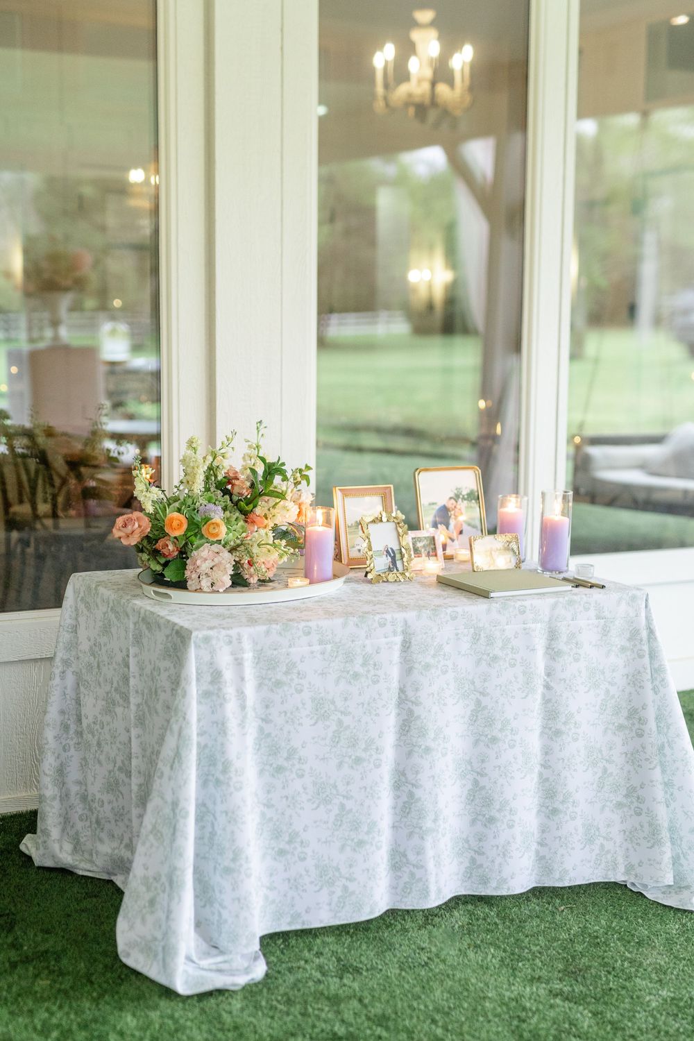 Wedding memory table with floral arrangement, framed photos, purple candles, and floral linen tablecloth.