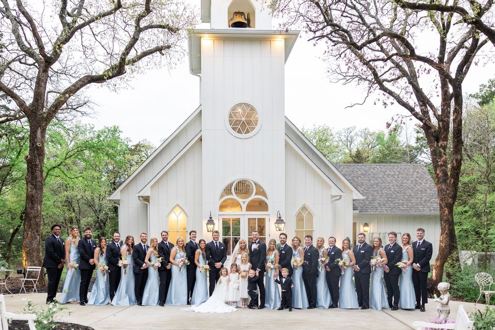 Large wedding party posing in front of a white chapel surrounded by trees in soft blue and gray attire.