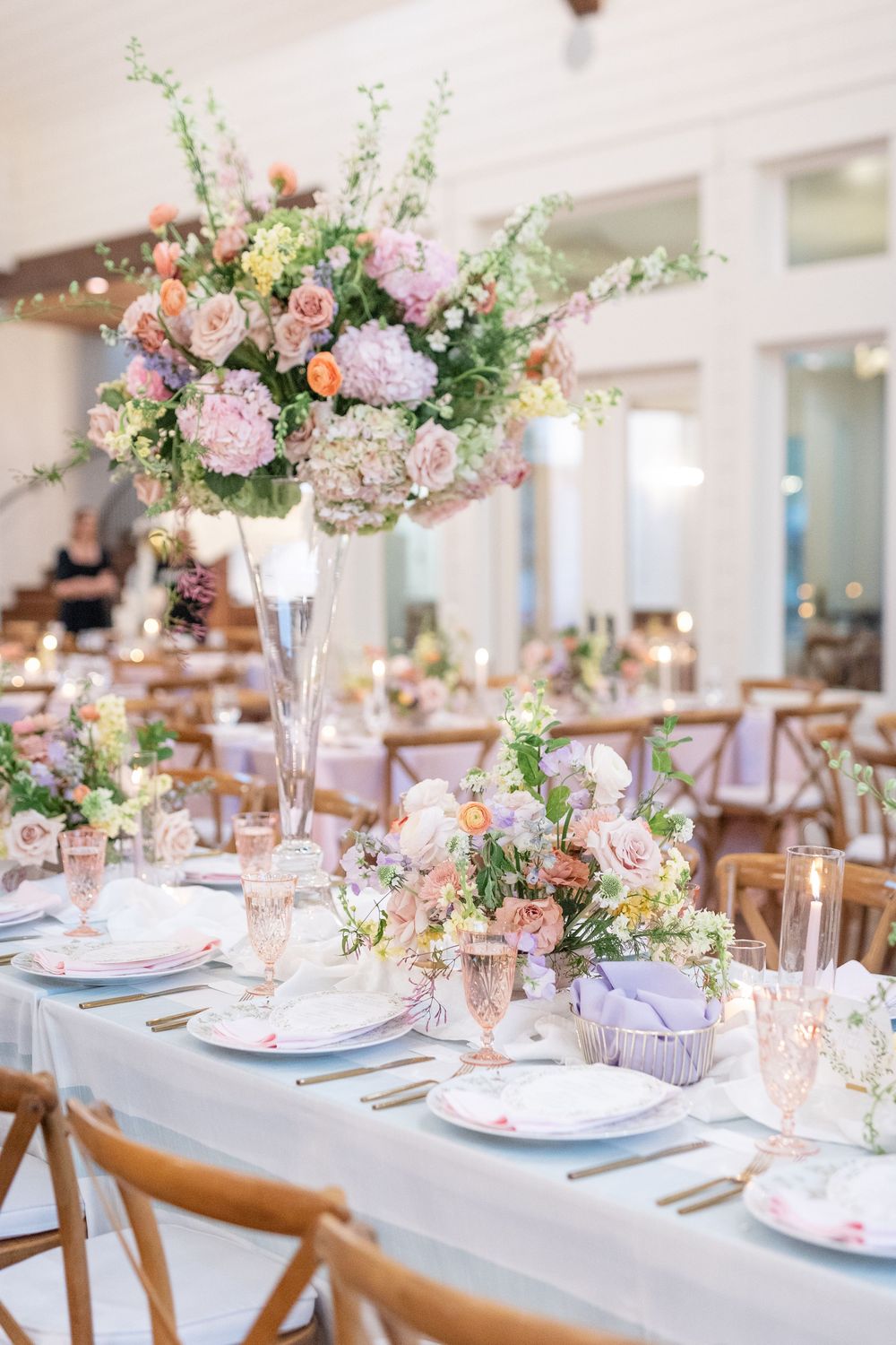 Elegant reception tables with tall silver vases of pastel blooms and pink crystal glassware on blue linens.