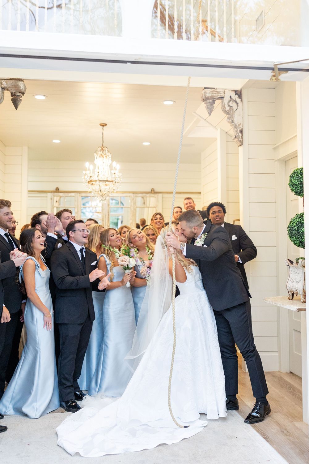 Bride and groom share first kiss at altar surrounded by cheering wedding party in elegant white venue.