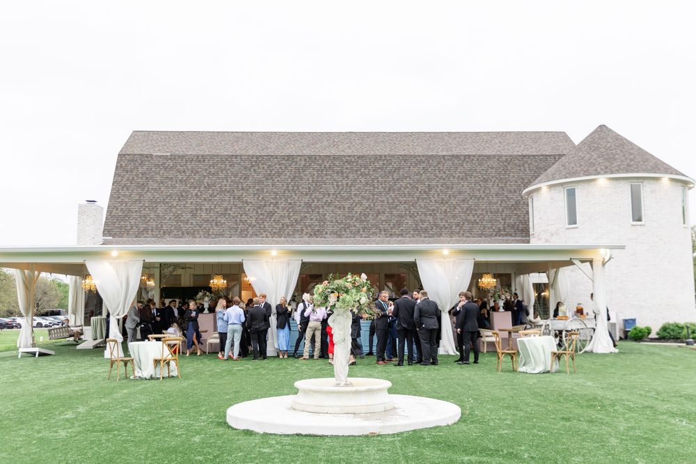 Guests mingle on green lawn at elegant white wedding venue with covered pavilion and fountain centerpiece.