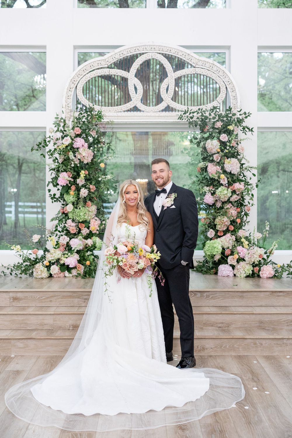 Bride and groom pose before floral arch with pink blooms and greenery in bright indoor ceremony space.
