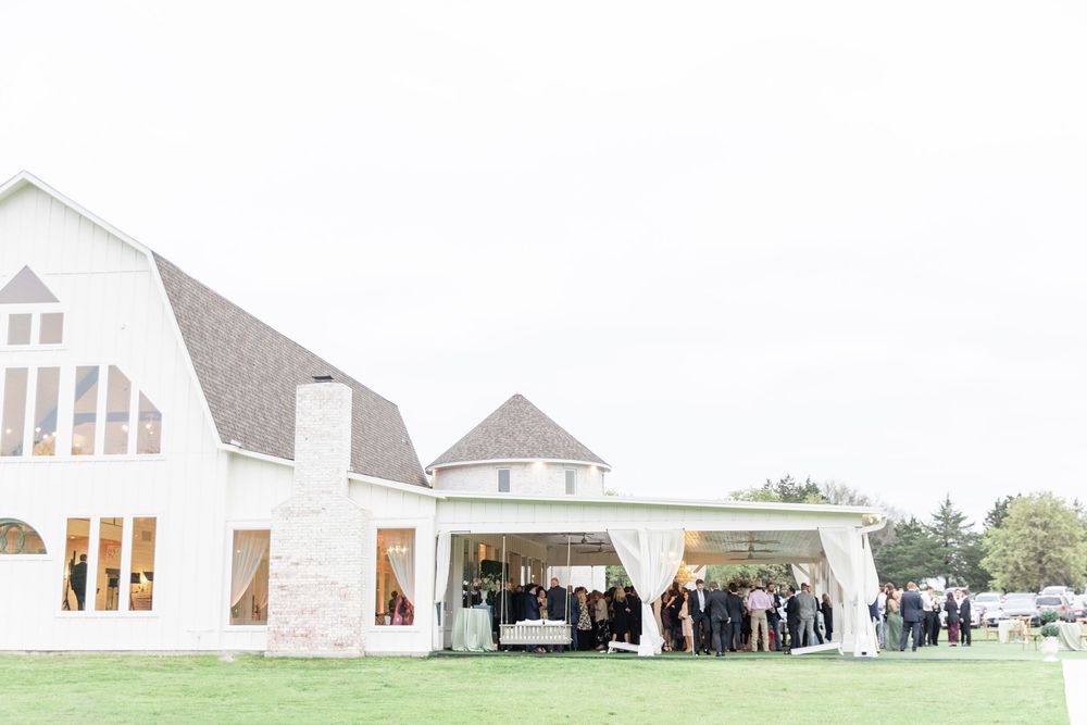 Aerial view of wedding guests gathered under draped pavilion beside white barn-style wedding venue.