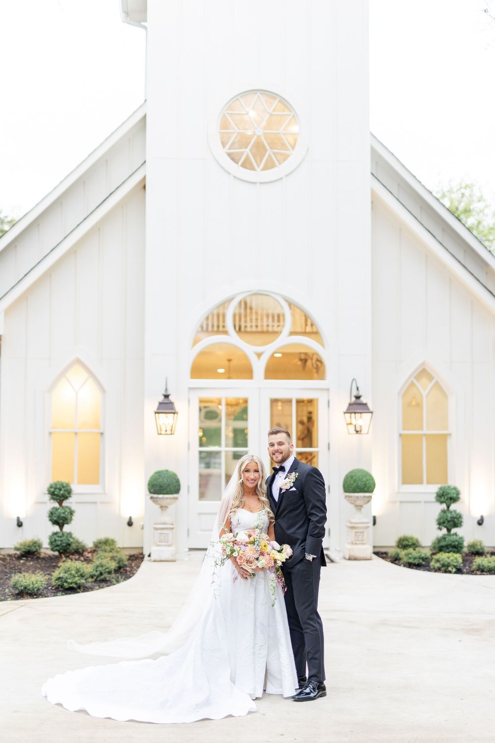 Bride and groom portrait in front of white chapel wedding venue with Gothic arched windows at dusk.
