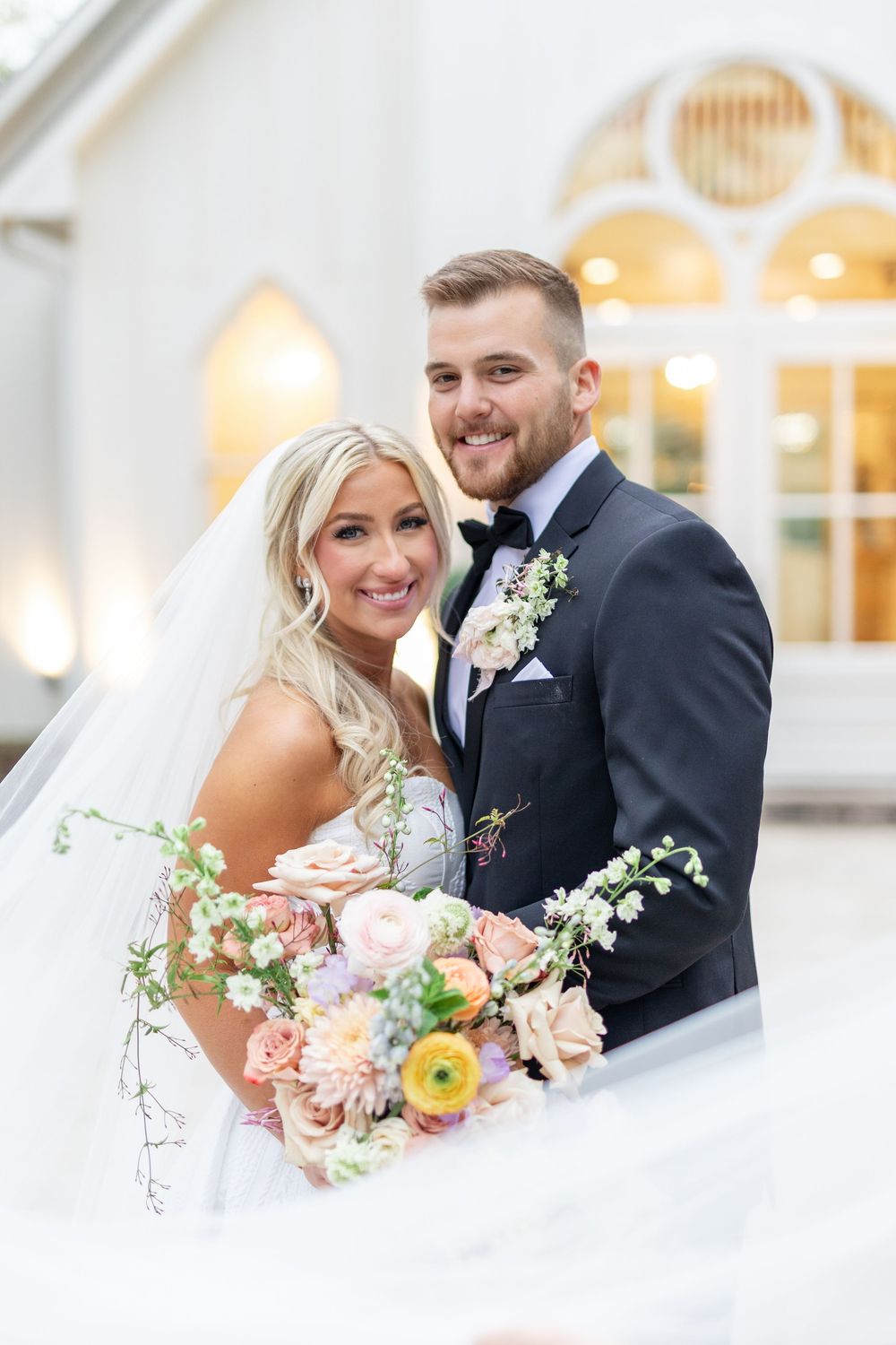 Smiling bride and groom pose with colorful peach and pink bridal bouquet outside white wedding chapel.