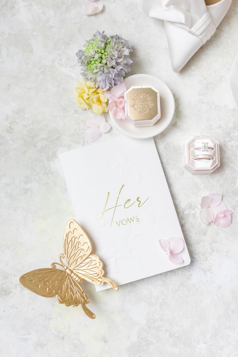 Elegant bridal flat lay with Her Vows book, gold butterfly, diamond rings in pink velvet box, pearl necklace, and pastel flowers.