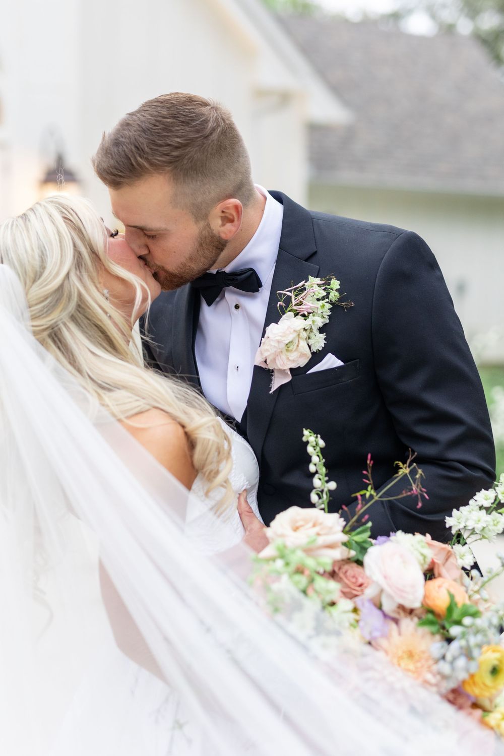 Bride and groom share romantic kiss beneath flowing veil with lush colorful bridal bouquet in foreground.