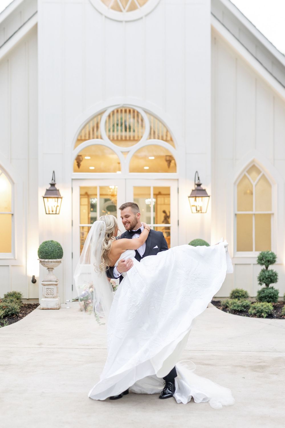 Groom lifts bride in flowing white gown outside elegant white chapel wedding venue at golden hour.