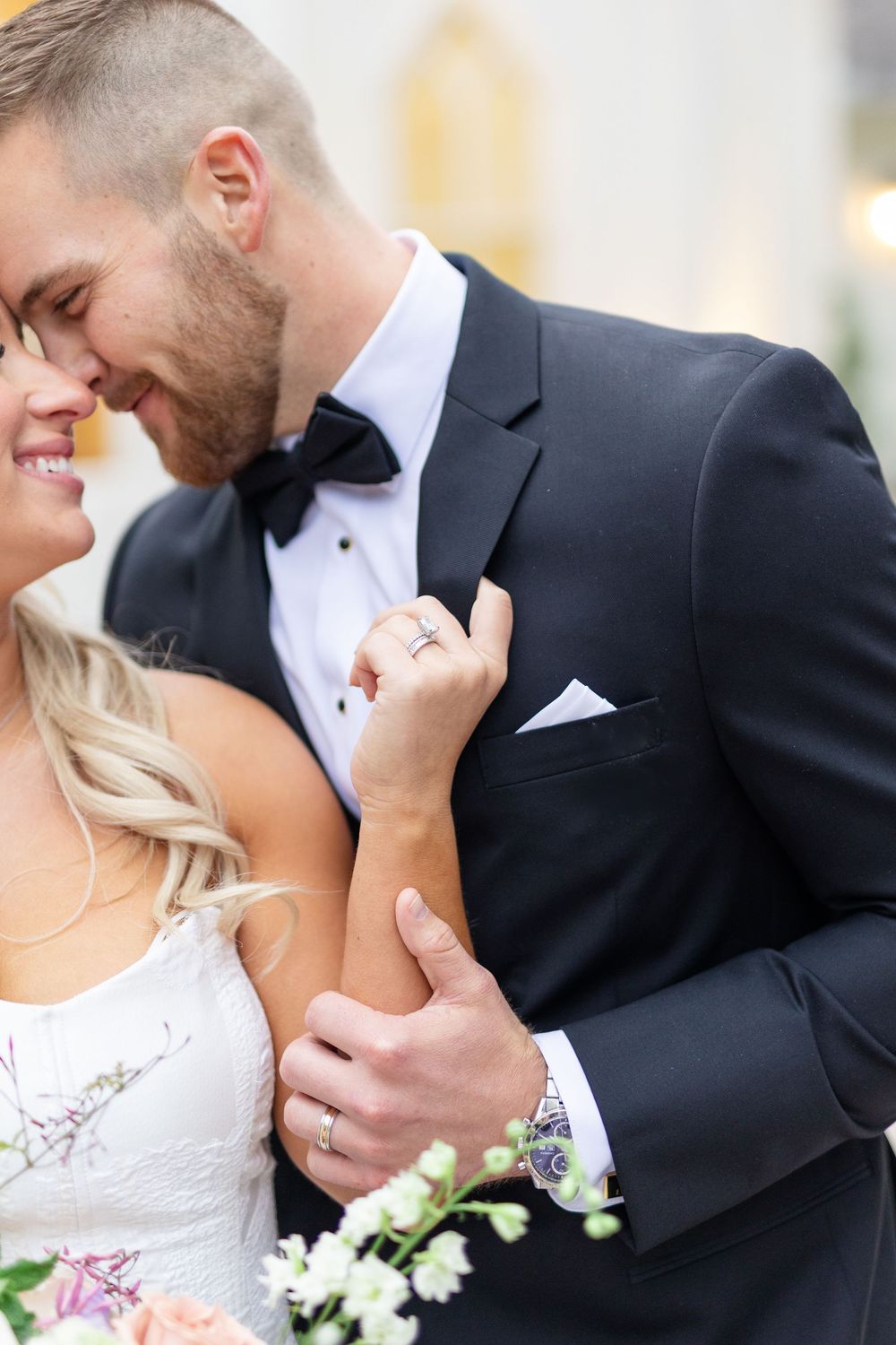 Groom nuzzles smiling bride as she holds his lapel wearing diamond wedding ring outside white venue.