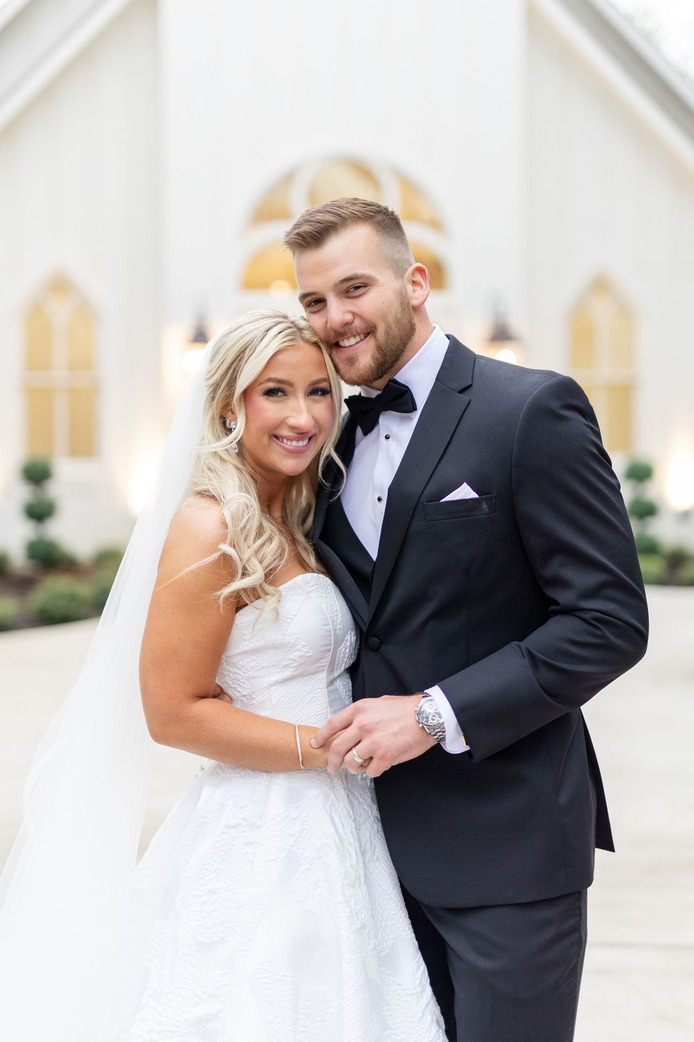 Smiling bride and groom pose together in front of white Gothic-style chapel wedding venue exterior.