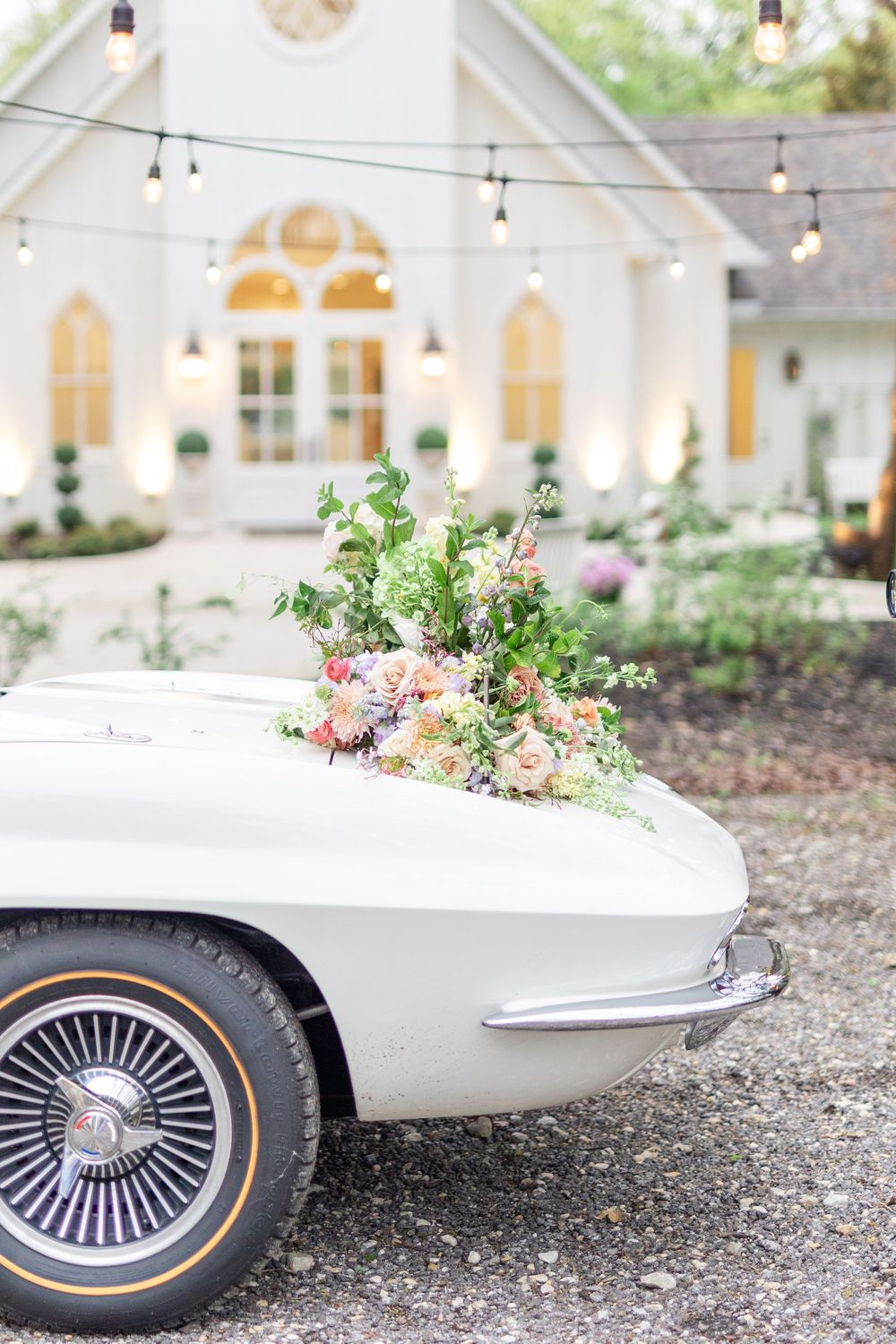 Vintage white Corvette adorned with lush floral arrangement parked outside glowing white wedding chapel.