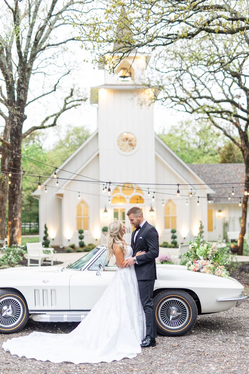 Bride and groom embrace in front of a white chapel and vintage Corvette Stingray at an elegant outdoor wedding.