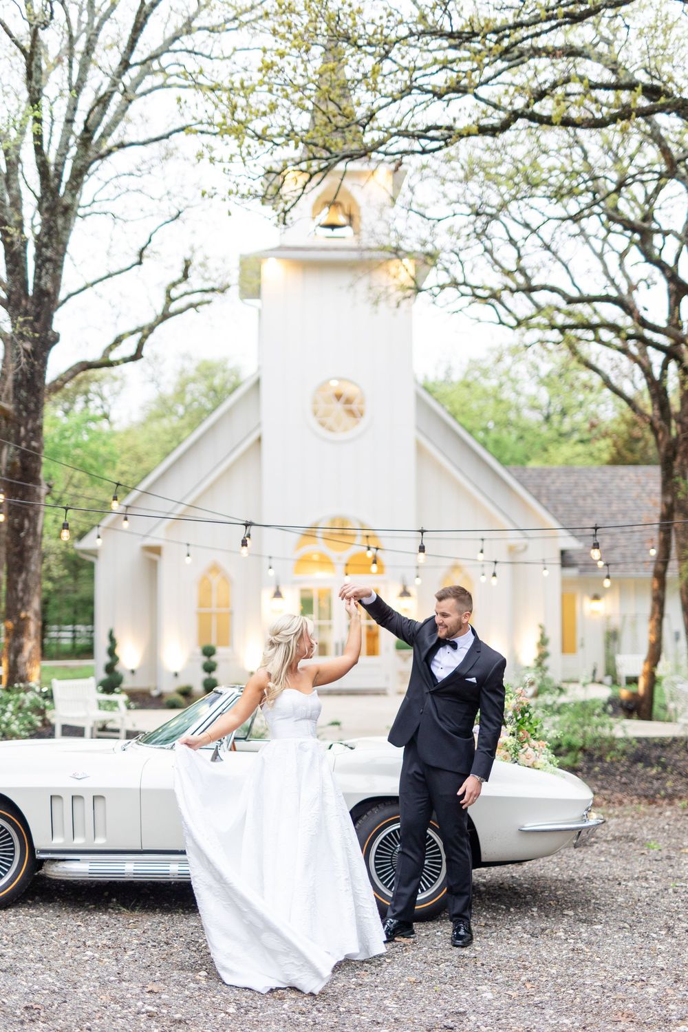 Groom twirls bride beside a classic white Corvette in front of a charming white wedding chapel with string lights.
