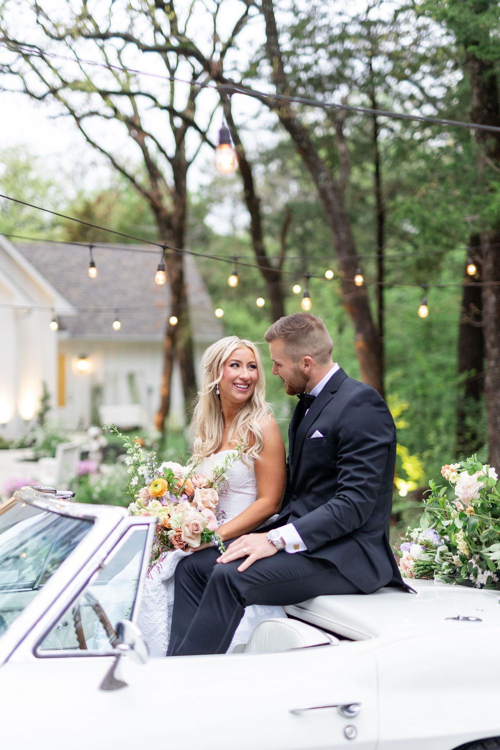 Bride and groom sit together on a vintage white Corvette adorned with pastel floral arrangements under string lights.