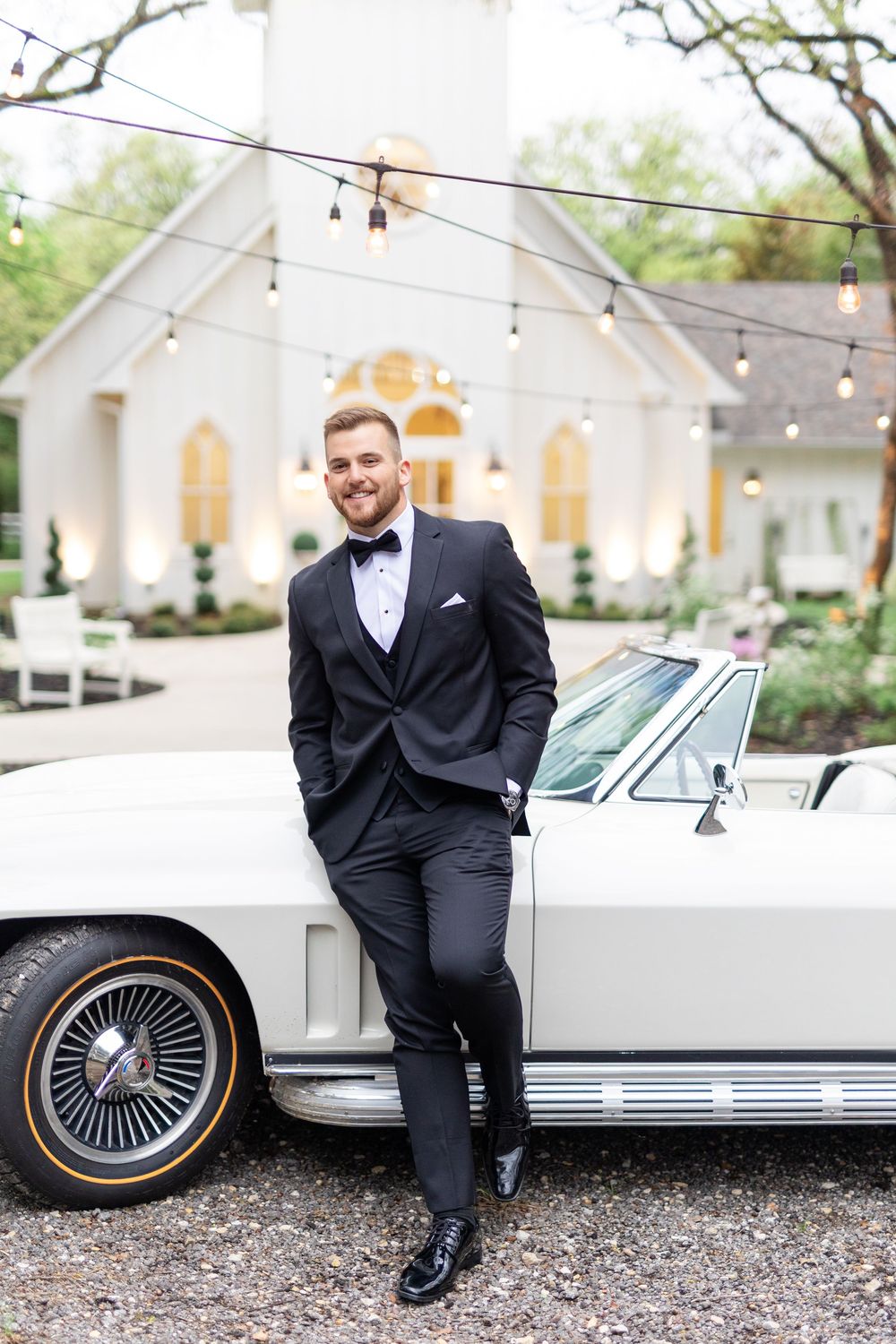 Handsome groom in black tuxedo leans against a vintage white Corvette in front of a white wedding chapel.