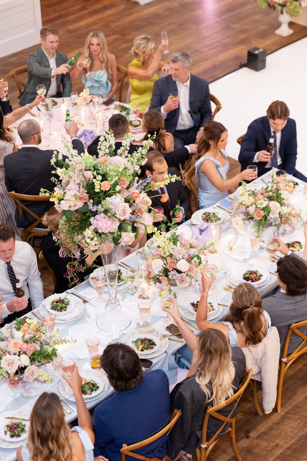 Wedding guests raise glasses in a toast at a beautifully decorated reception table with tall floral centerpieces.