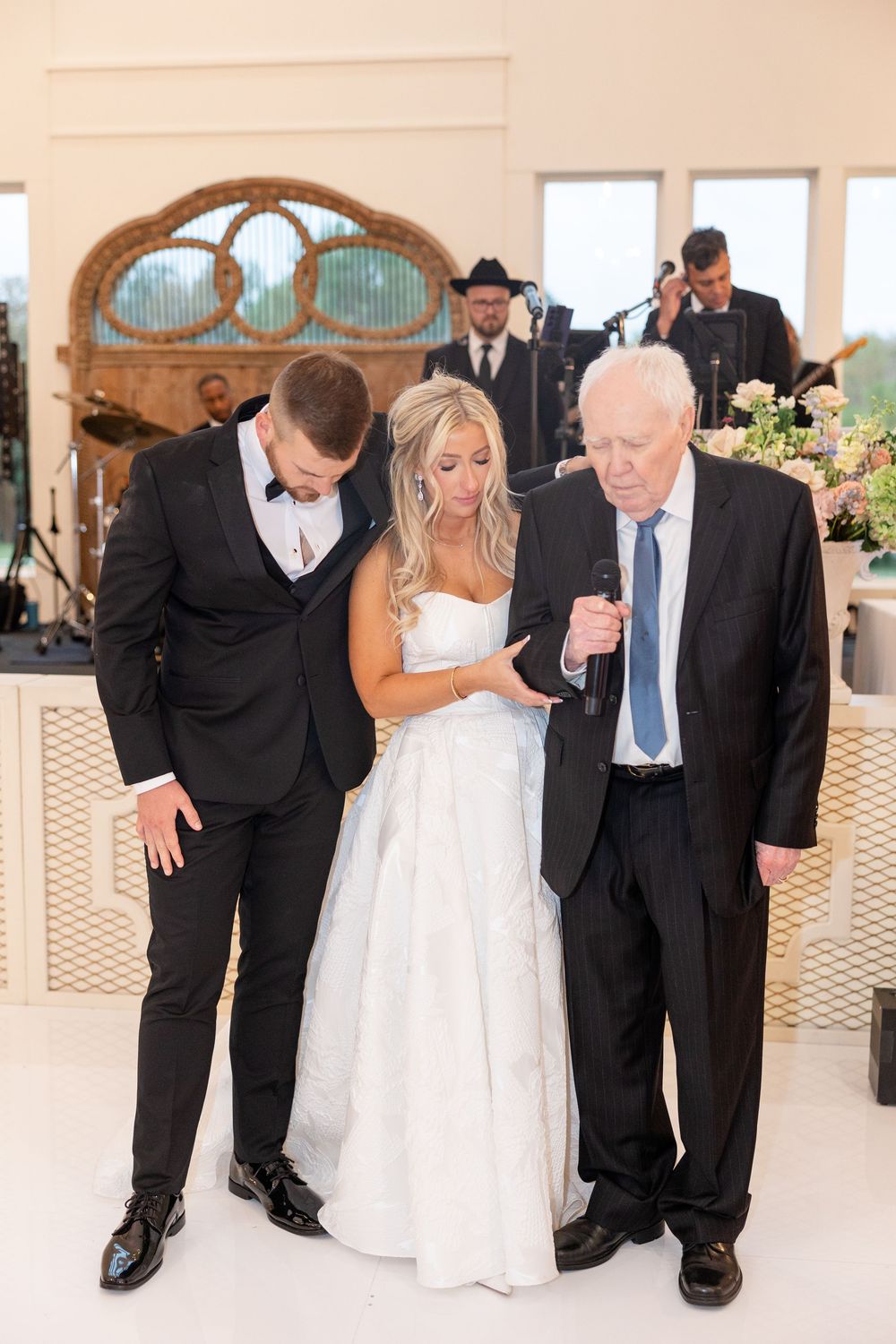 Elderly officiant prays with bride and groom on the dance floor during an elegant indoor wedding reception.