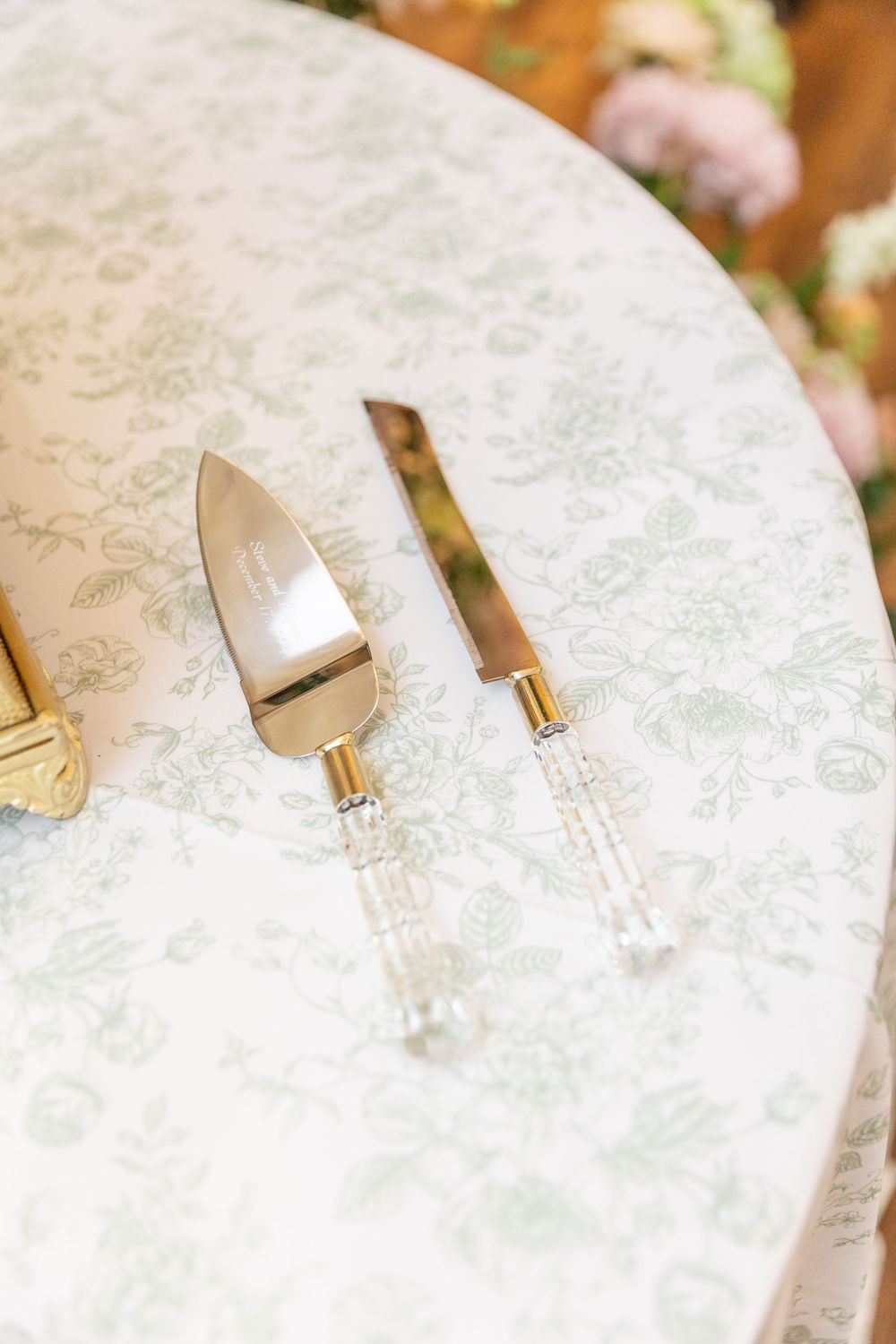 Gold and crystal wedding cake serving set displayed on a floral patterned tablecloth at an elegant reception.