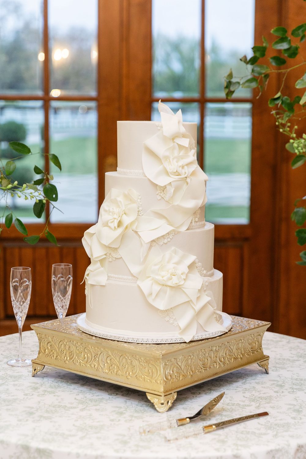 Four-tier ivory wedding cake adorned with sugar ruffles on a gold stand beside crystal champagne flutes.
