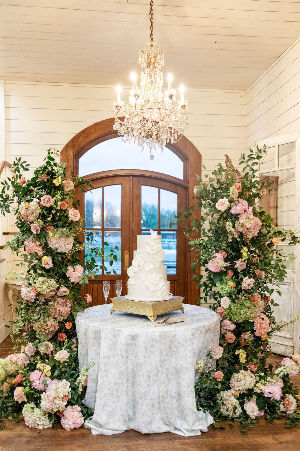 Elegant wedding cake display with floral arch of pink roses and greenery framing arched wooden doors with chandelier.