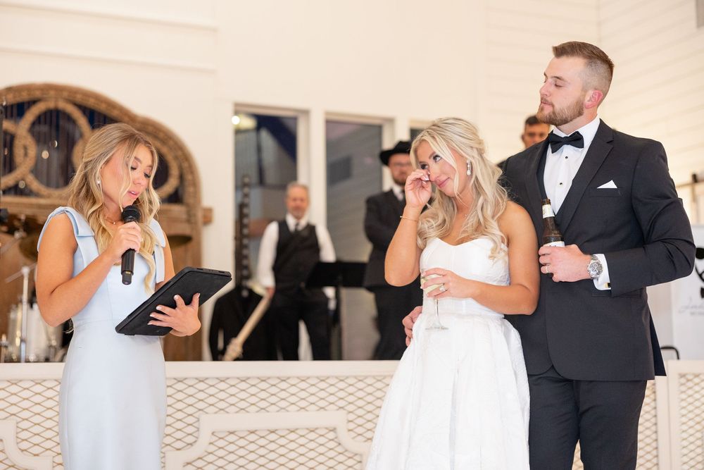 Bride wipes tears during heartfelt wedding reception speech by bridesmaid while groom stands proudly beside her.