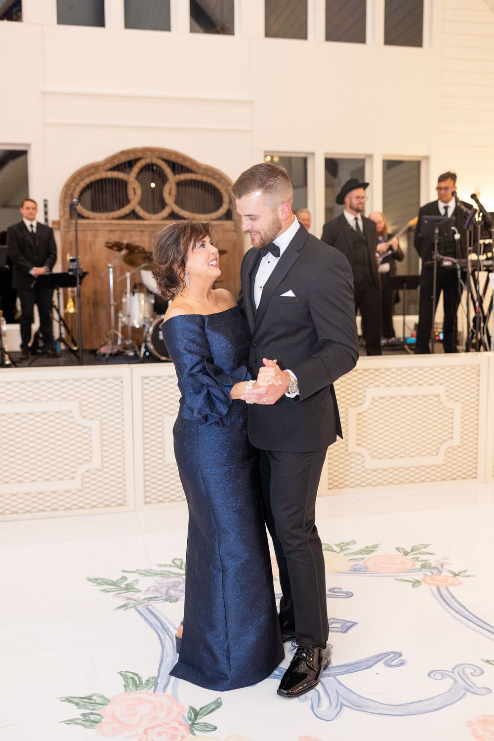 Groom shares tender mother-son dance on floral monogram dance floor with live band performing in background.