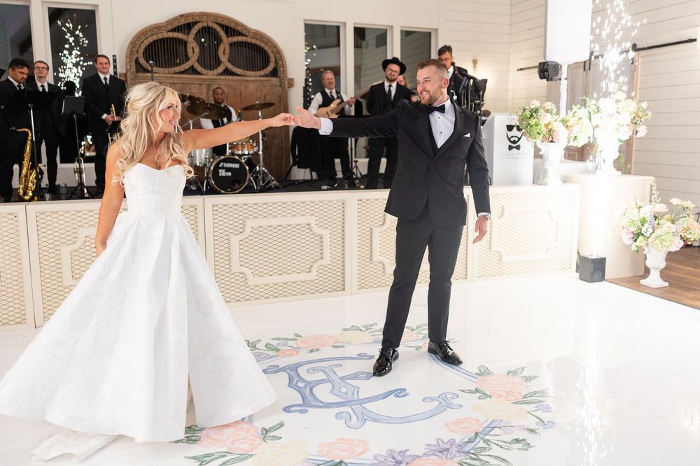 Bride and groom spin joyfully during first dance on floral monogram floor with live band performing.