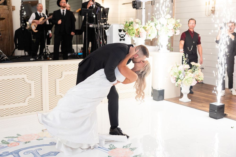 Groom dips bride for romantic kiss on floral monogram dance floor with cold sparkler effects at reception.
