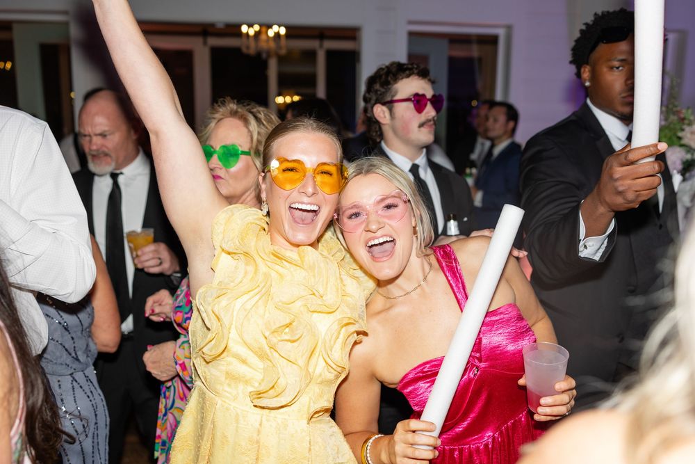 Two women in colorful dresses and orange sunglasses celebrate joyfully at a wedding reception party.