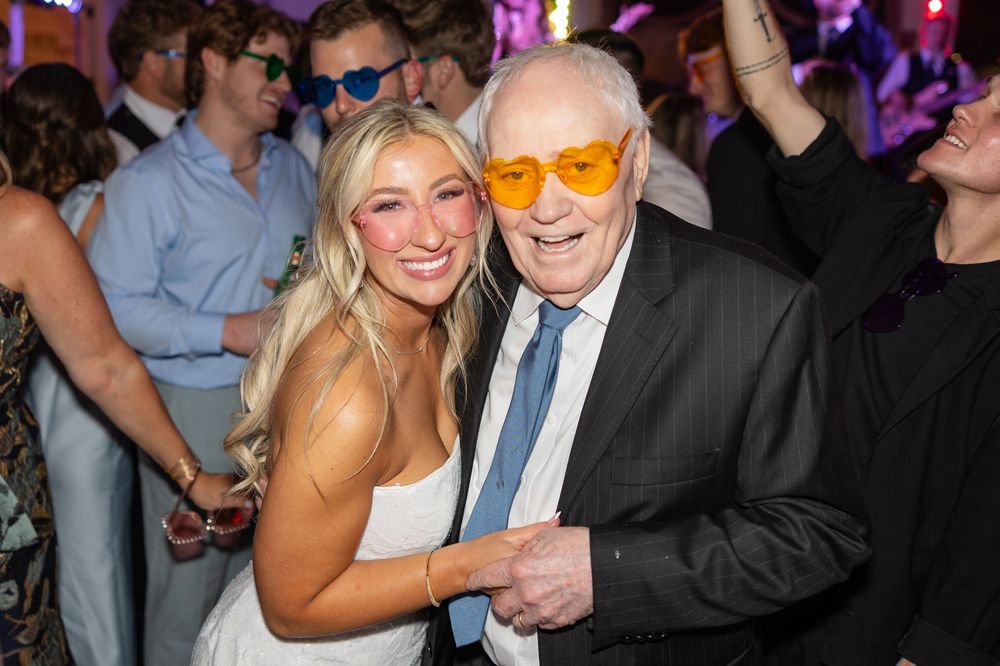 Young woman poses with elderly man wearing orange glasses at a festive wedding reception dance floor.