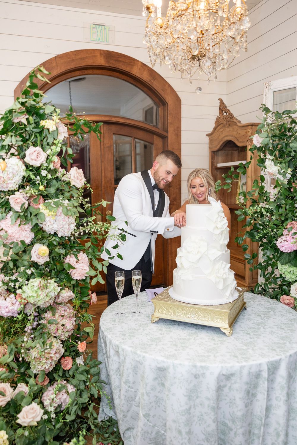 Bride and groom cut elegant white tiered wedding cake surrounded by lush floral arrangements and chandelier.