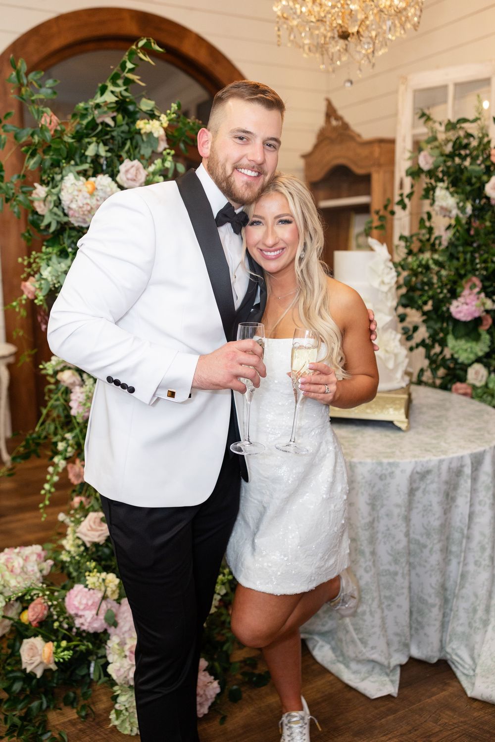 Bride and groom pose with champagne glasses beside floral arrangements and wedding cake in elegant venue.