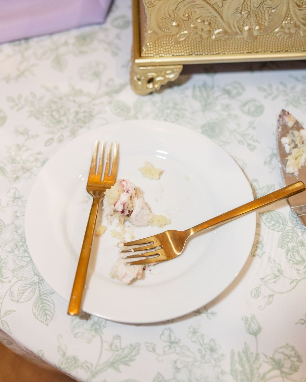 Two gold forks rest on white plate with wedding cake remnants on floral tablecloth after reception.