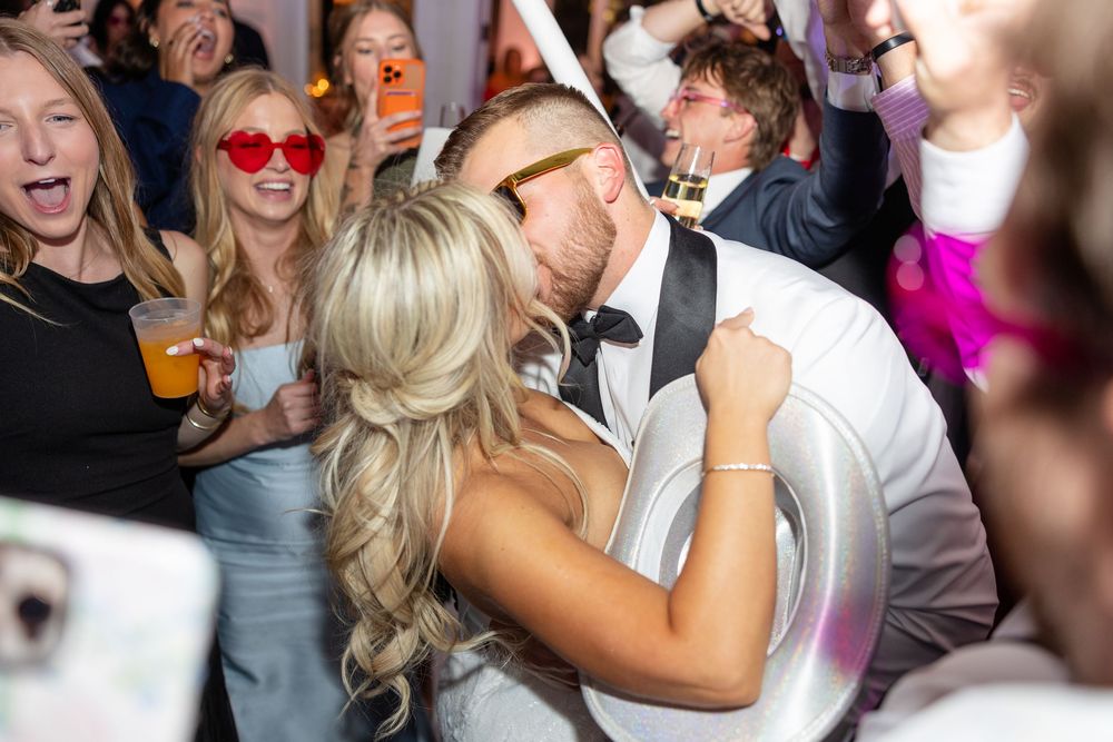 Bride and groom share passionate kiss surrounded by cheering guests on the wedding reception dance floor.