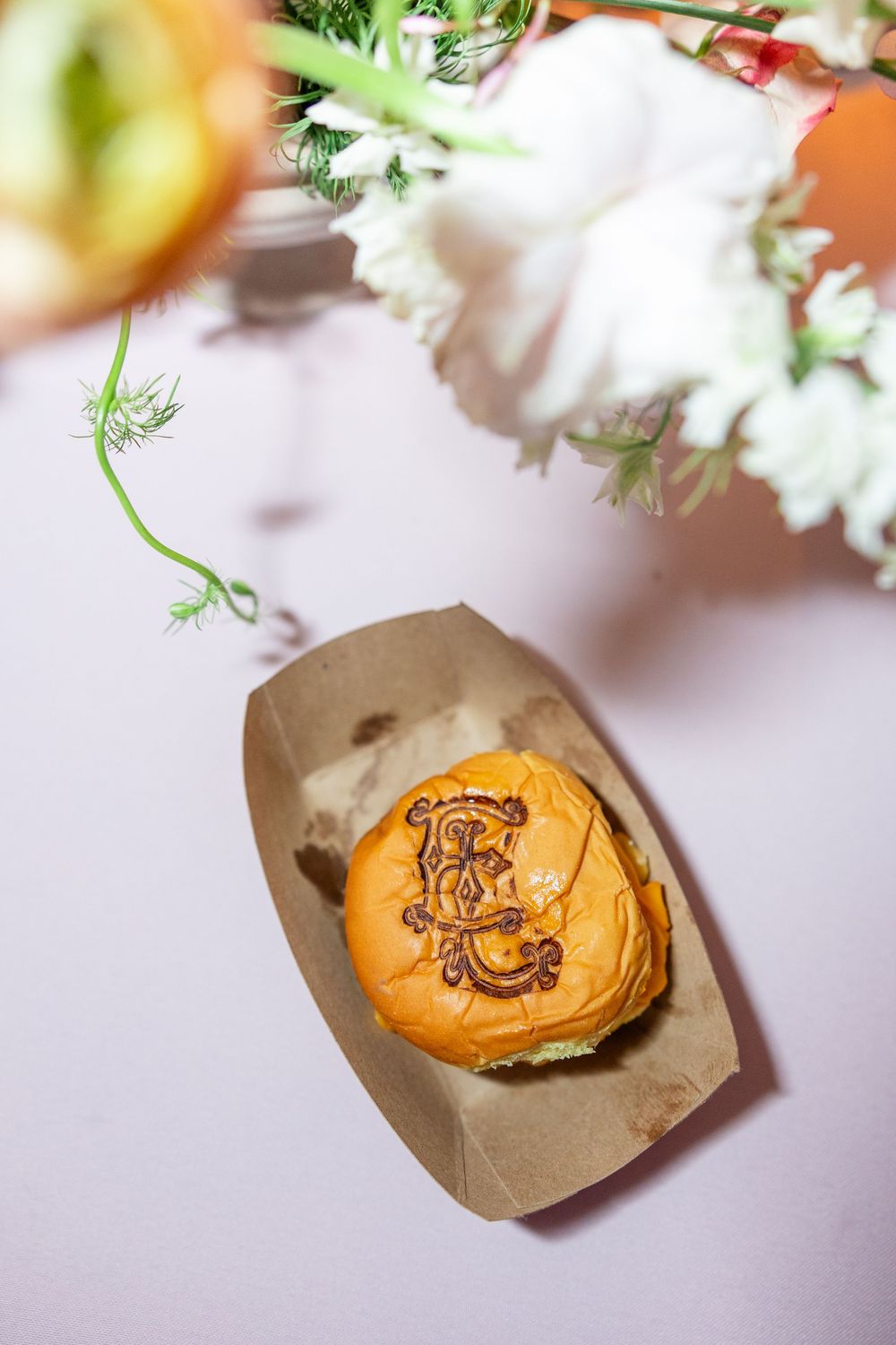 Custom monogram-branded slider burger served in kraft paper boat beside white wedding flowers centerpiece.
